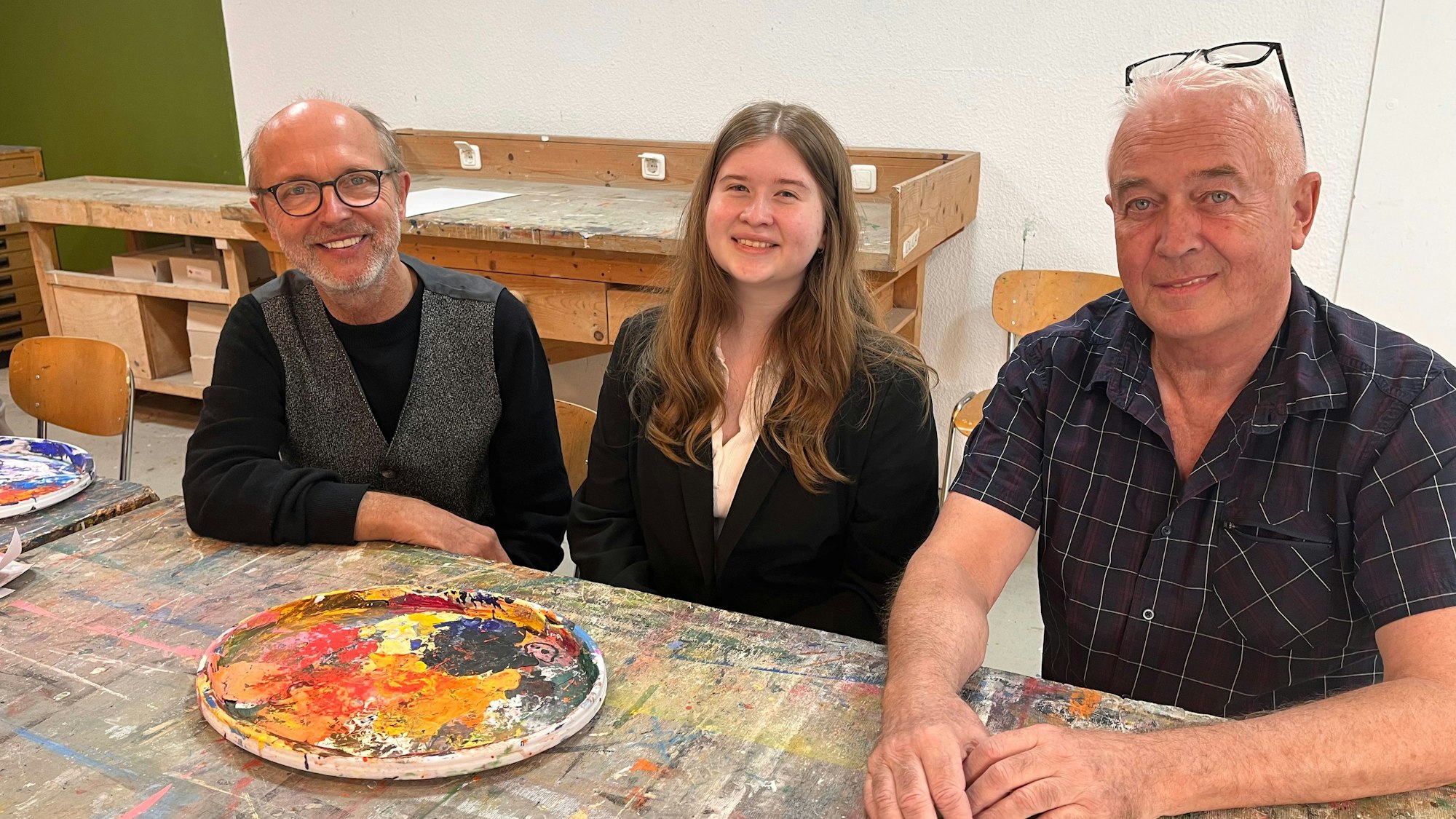 Zwei Männer und eine Frau sitzen an einem Tisch, auf dem Malfarben in einem runden Teller stehen.