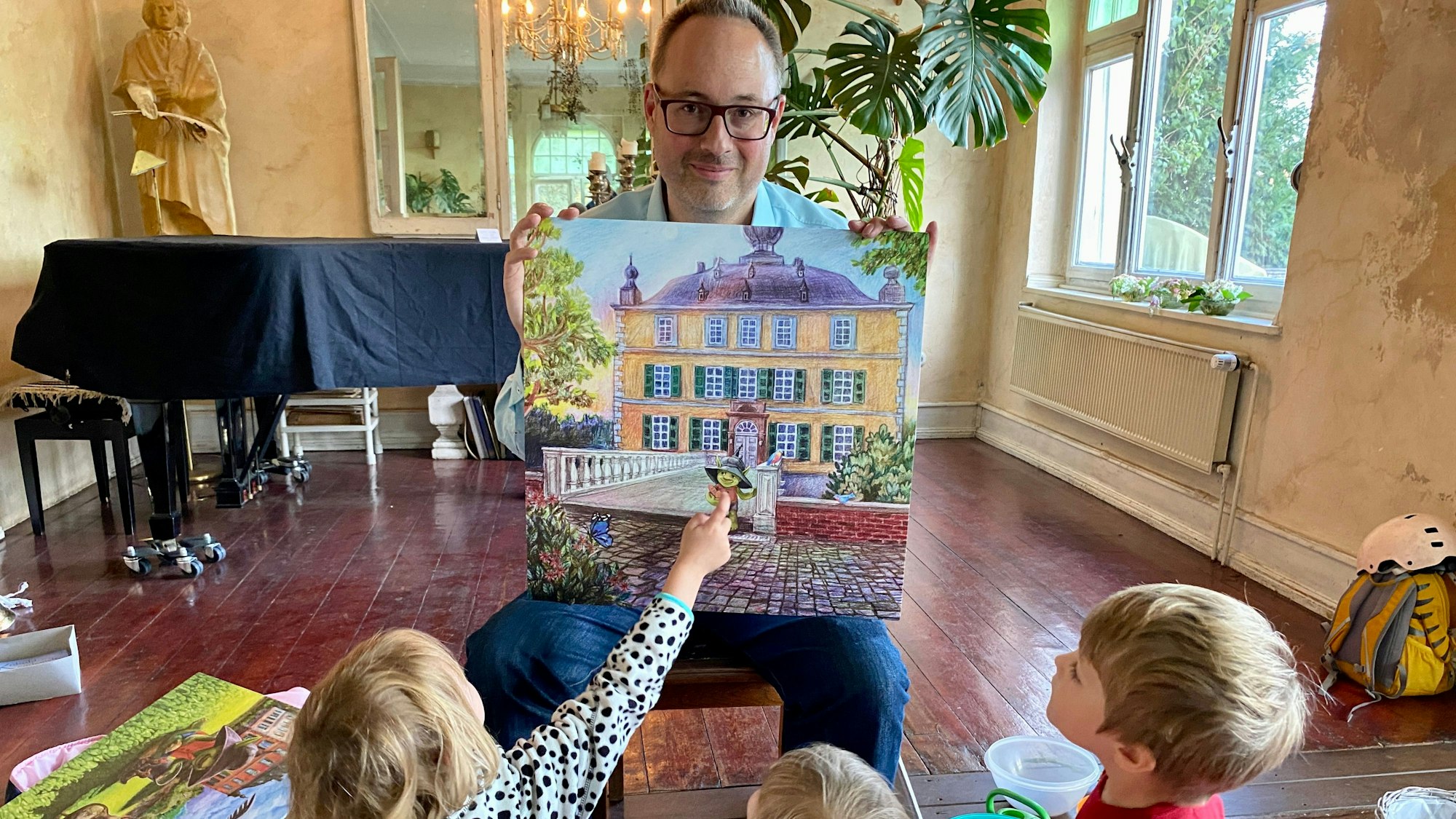 Der Autor hält eine Zeichnung in der Hand, auf der grüne Kobold vor einem prächtigen Gebäude zu sehen ist.