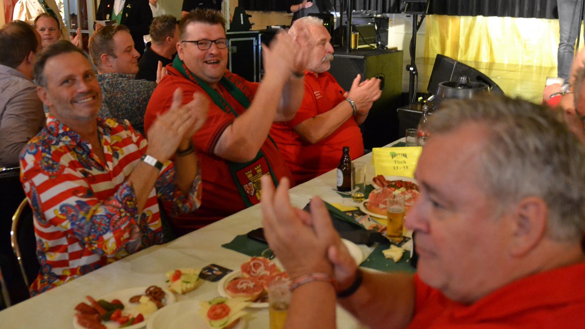 Einige Männer sitzen gut gelaunt am Tisch und klatschen zur Musik. Es gibt Bier und Mettbrötchen.