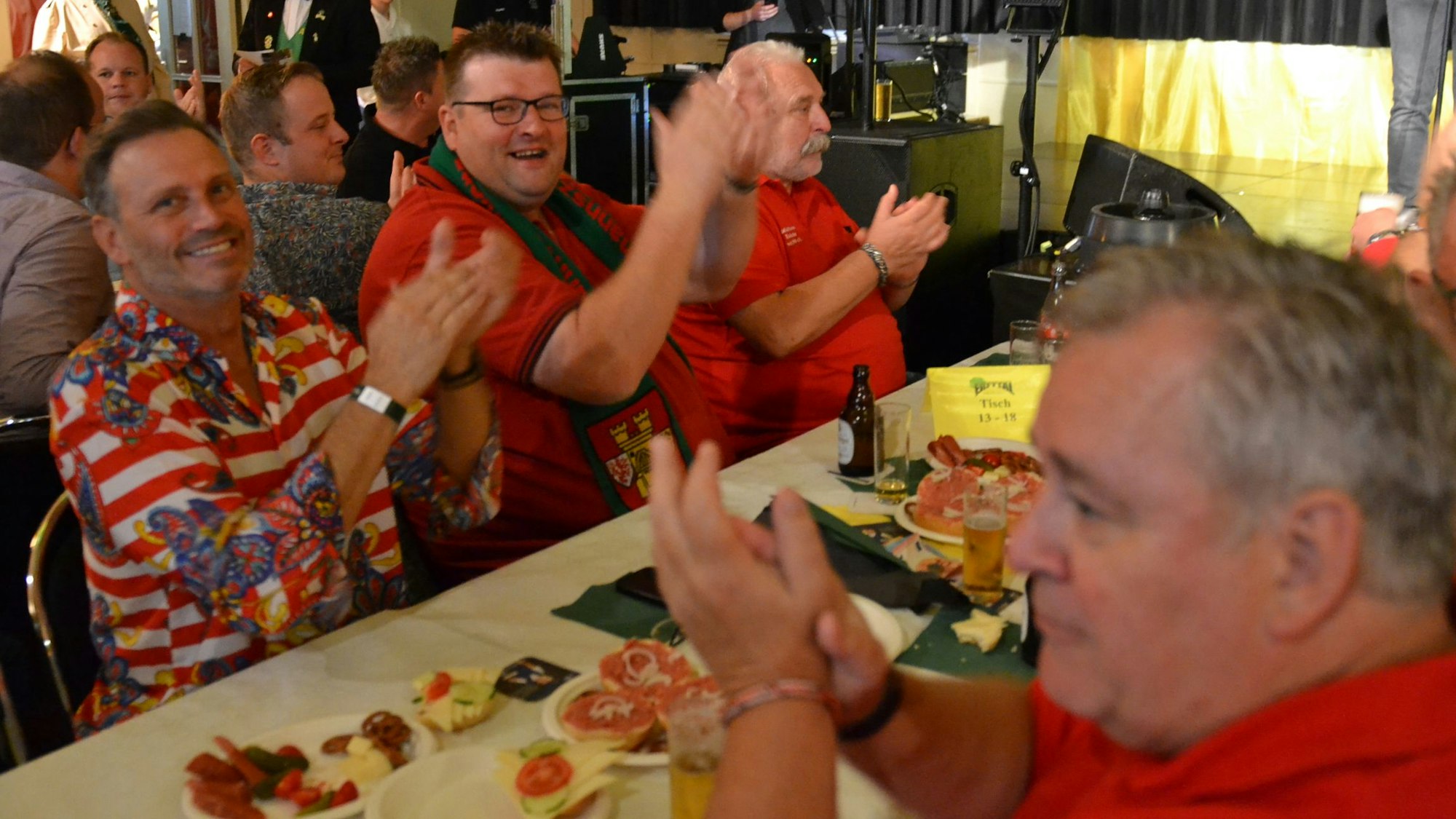 Einige Männer sitzen gut gelaunt am Tisch und klatschen zur Musik. Es gibt Bier und Mettbrötchen.