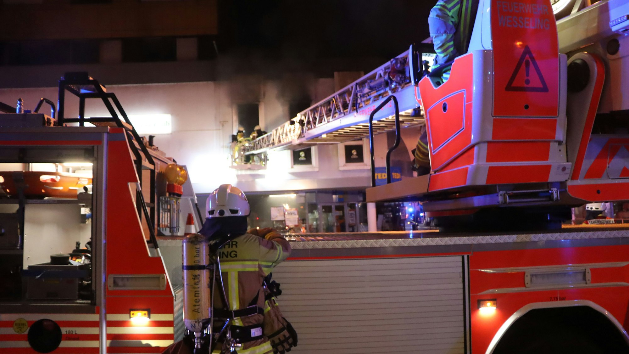 Zu sehen ist ein Feuerwehrwagen sowie im Hintergrund ein Haus, aus dessen Fenstern Rauch quillt.