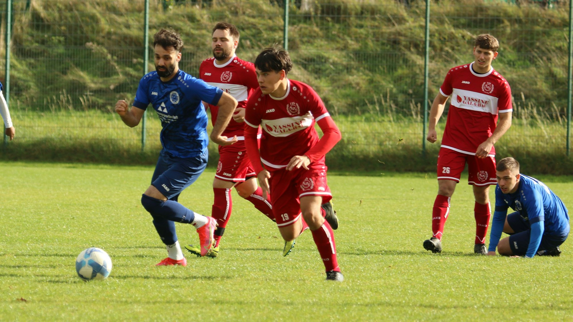 Die beiden Fußballer im Zweikampf. In Hintergrund kniet ein Fußballer auf dem Boden, ein anderer schaut dem Geschehen zu.