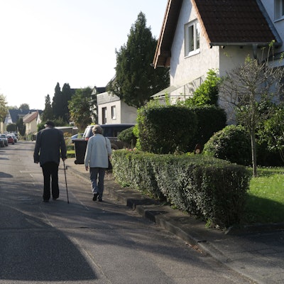 Ein älteres Paar geht auf der Fahrbahn einer Straße. Der Gehweg ist sehr schmal.