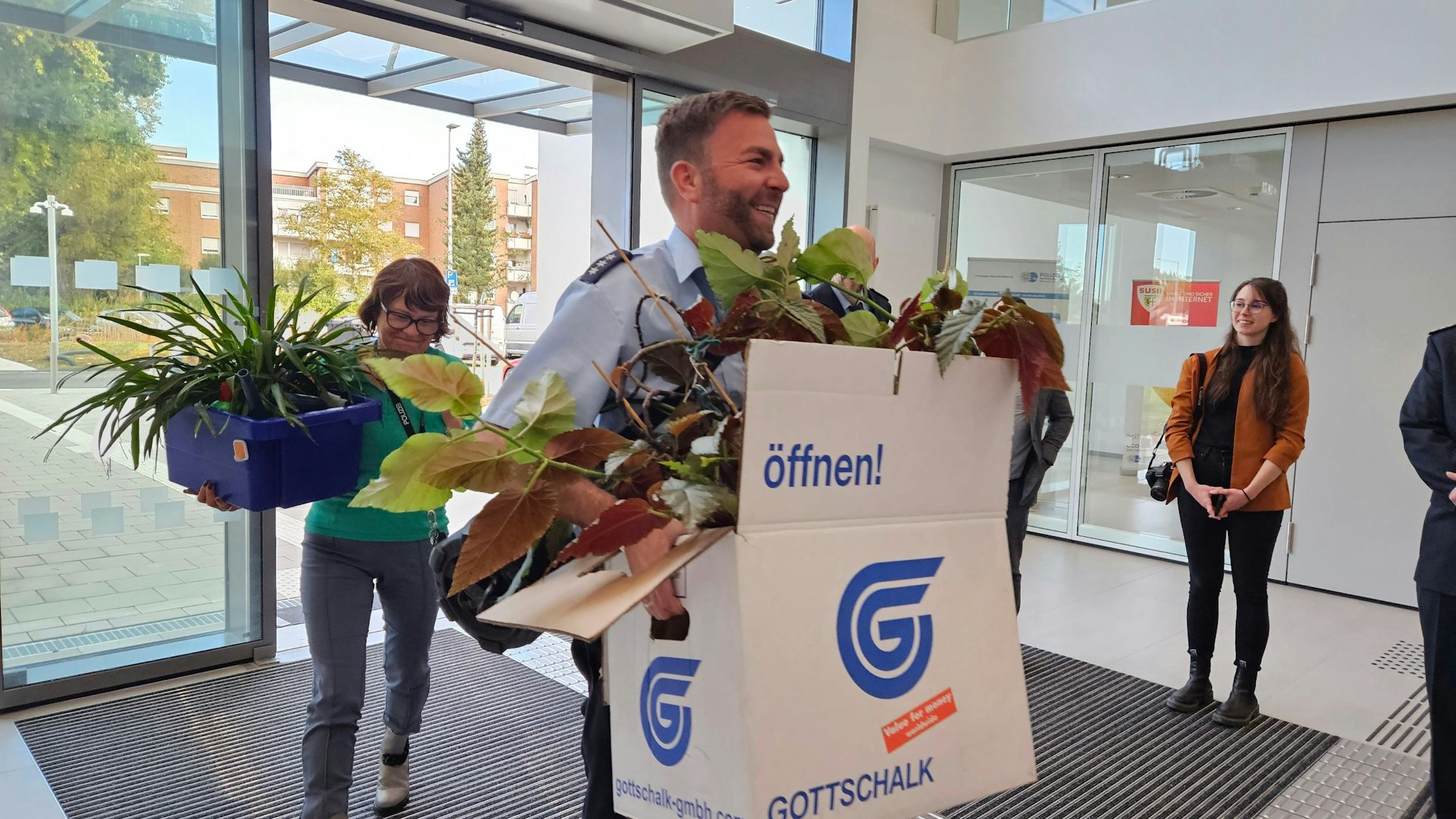 Ein Polizeibeamter trägt einen Umzugskarton mit Zimmerpflanzen in ein Gebäude, eine Frau, ebenfalls mit Kiste und Zimmerpflanze, folgt ihm.