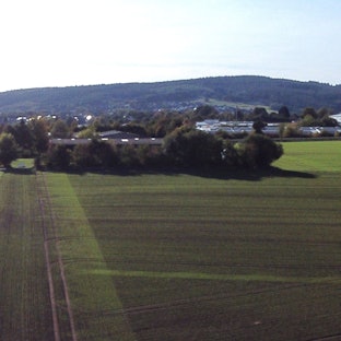 Zu sehen sind auf der Luftaufnahme der von Bäumen verdeckte Heidehof und die Freifläche daneben. Das Gelände wird von einer Straße begrenzt.