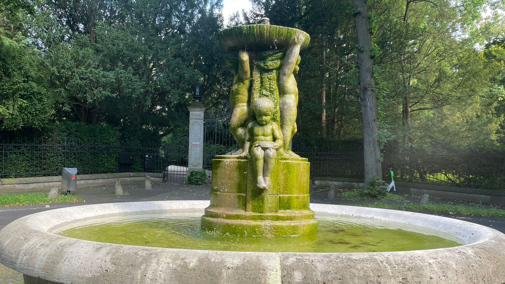 Ein steinerner Brunnen mit kindlichen Figuren steht in einer Parkanlage.