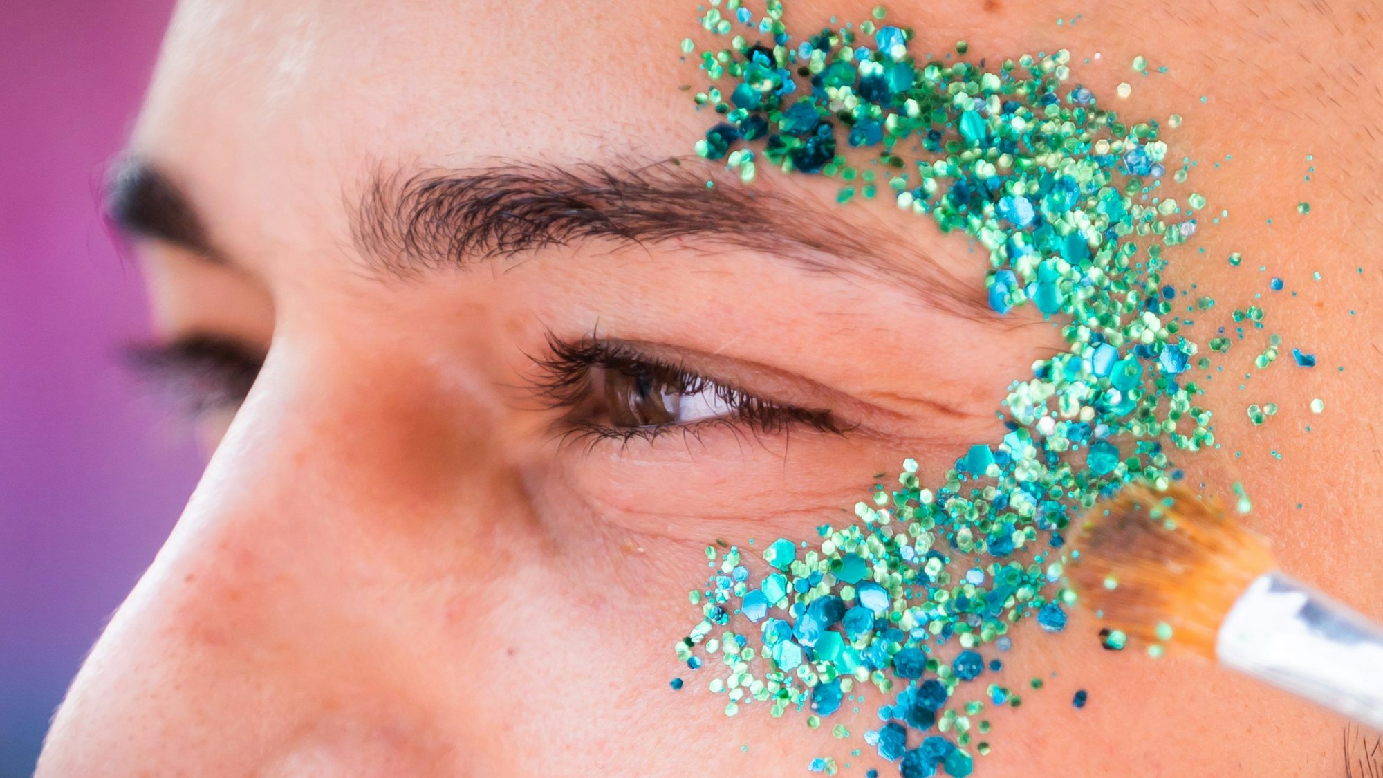 Ein Mann lässt sich mit Glitzer schminken: Ein Pinsel trägt türkise Partikel auf die Schläfe auf.