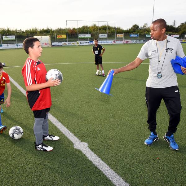 Abdoul Schwotzer Conde steht auf einem Fußballplatz und hält ein blaues Hütchen in der Hand, links von ihm steht ein Jugendlicher mit einem Fußball in den Händen.