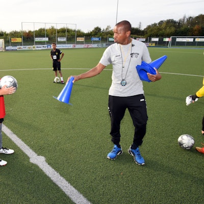 Abdoul Schwotzer Conde steht auf einem Fußballplatz und hält ein blaues Hütchen in der Hand, links von ihm steht ein Jugendlicher mit einem Fußball in den Händen.