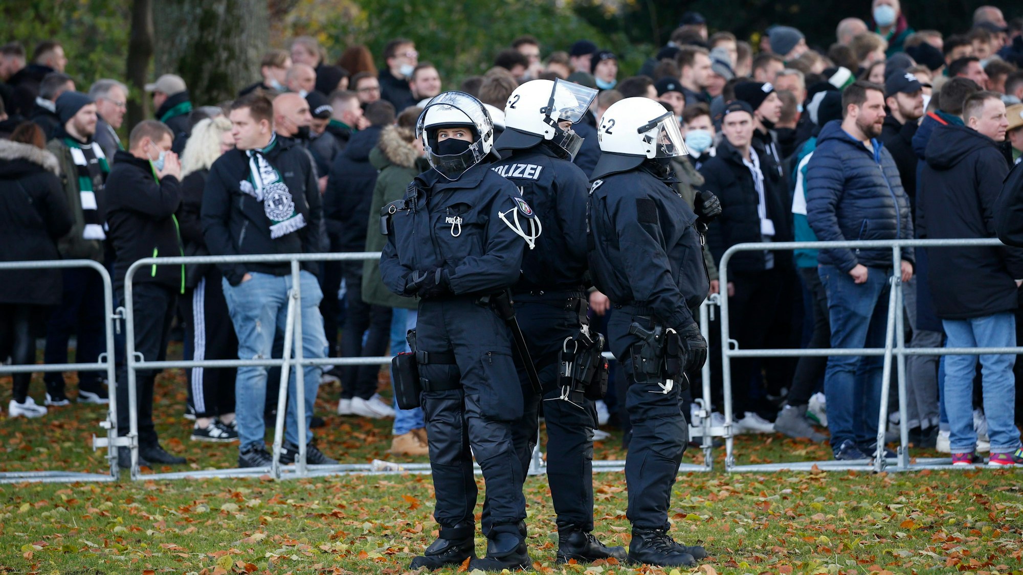 Polizisten stehen auf einer Wiese, dahinter stehen Fußballfans an einem Absperrgitter.
