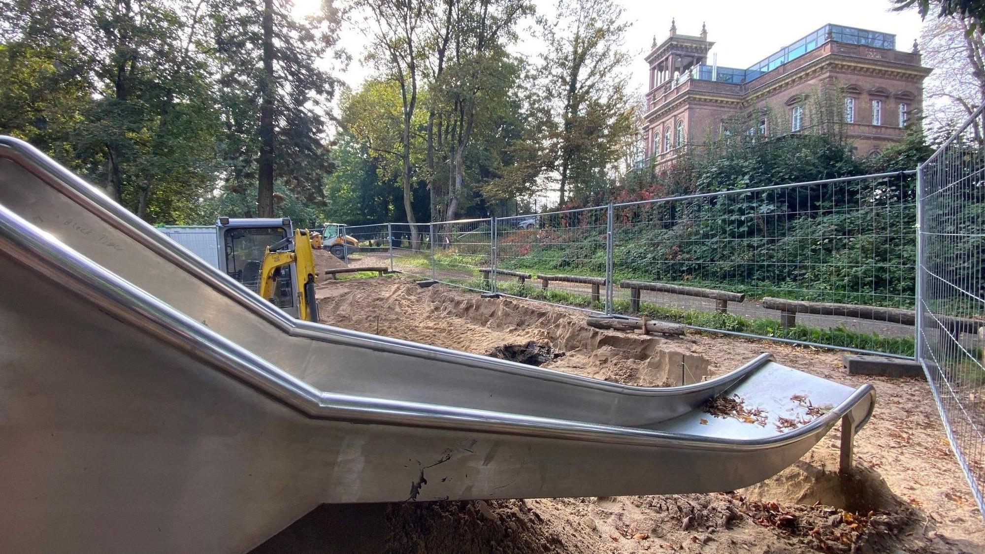 Zu sehen ist der Spielplatz mit einer silbernen Rutsche und im Hintergrund der historische Bahnhofsbau.