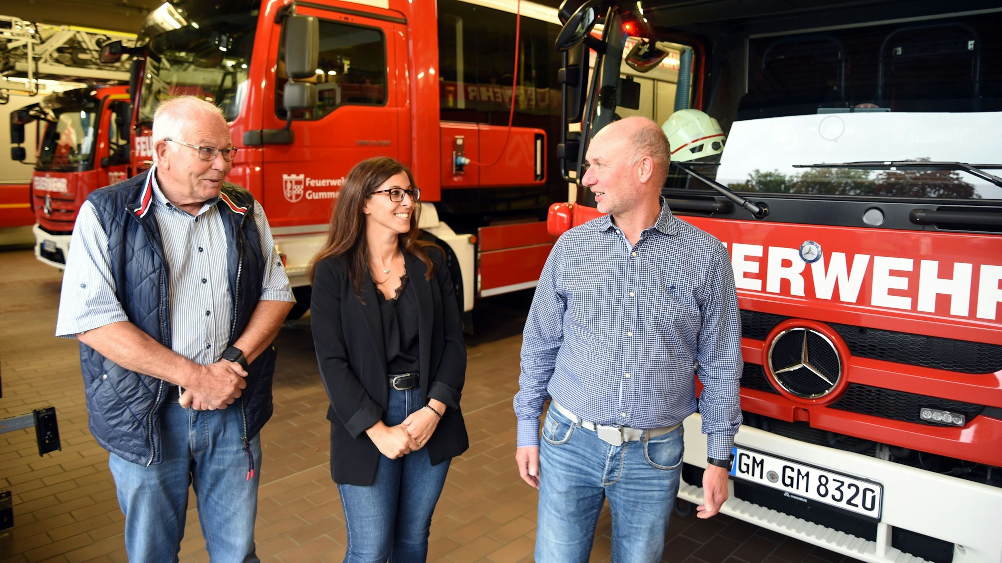Eine Frau und zwei Männer stehen vor Feuerwehrfahrzeugen.