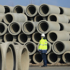 Zu einer hohen Wand aufgestapelte Kanalrohre. Davor steht ein Bauarbeiter mit einer gelb reflektierenden Jacke und einem Helm auf dem Kopf.