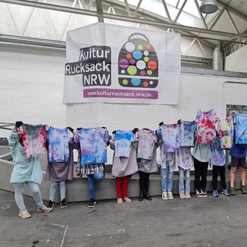 Jugendliche halten Batik-Shirts hoch, ihre Köpfe sind dahinter nicht zu sehen.