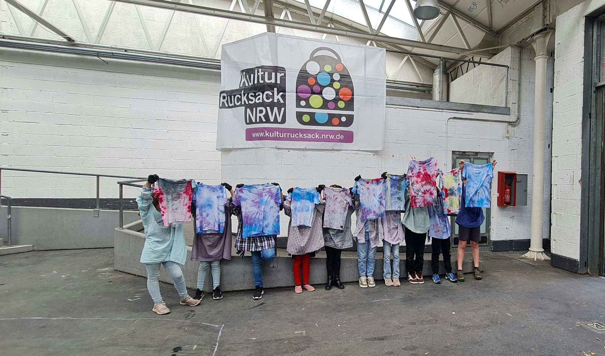 Jugendliche halten Batik-Shirts hoch, ihre Köpfe sind dahinter nicht zu sehen.