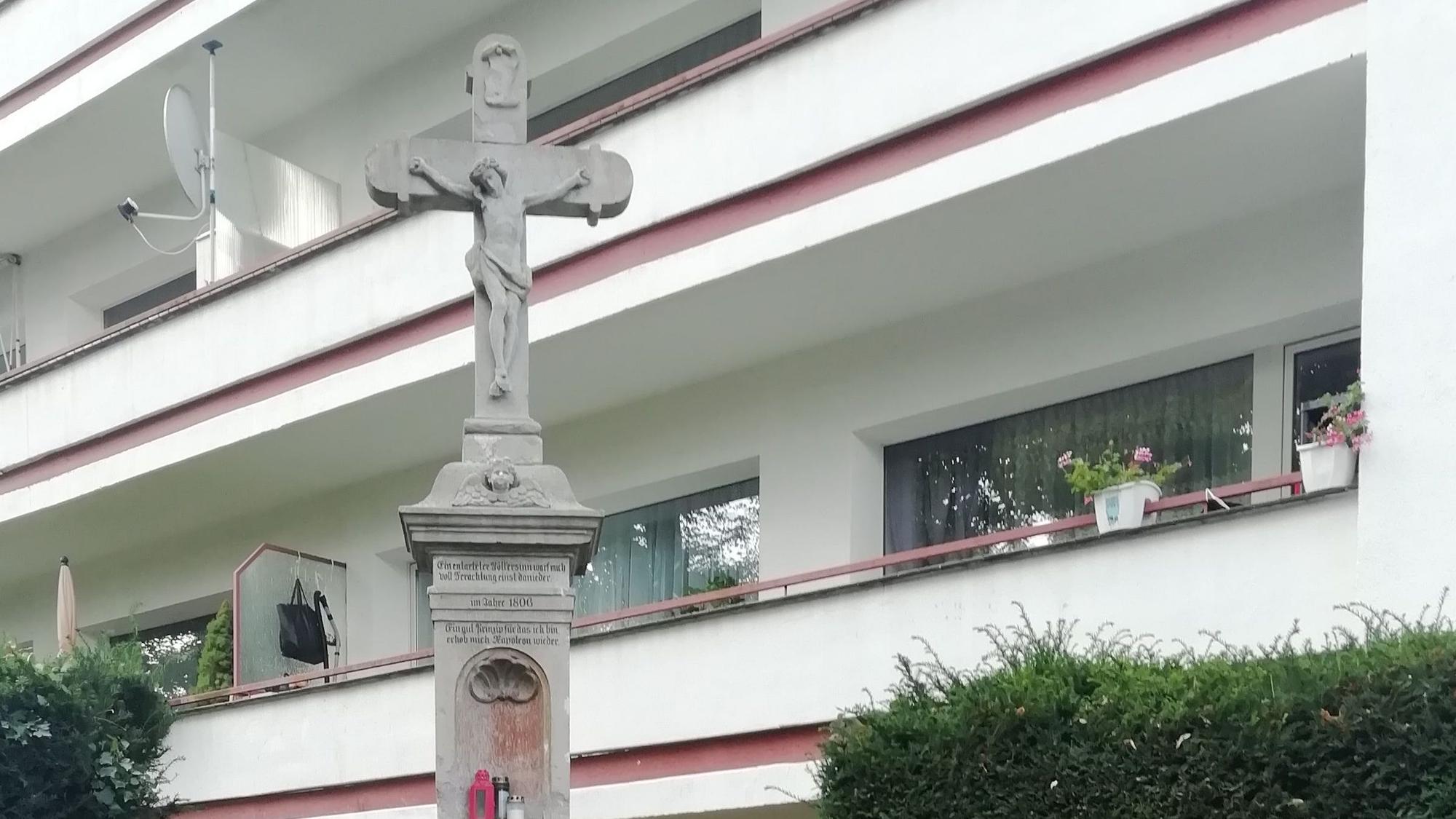 Ein steinernes Wegekreuz in einer Grünanlage vor einem Mehrfamilienhaus.