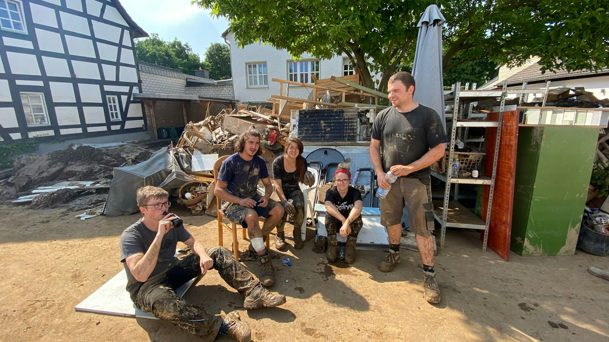 In Schweinheim machen Freiwillige, die nach der Flut beim Aufräumen helfen, eine Pause. Sie sitzen und stehen in der Nähe eines Fachwerkhauses, zum Teil mit Getränken.