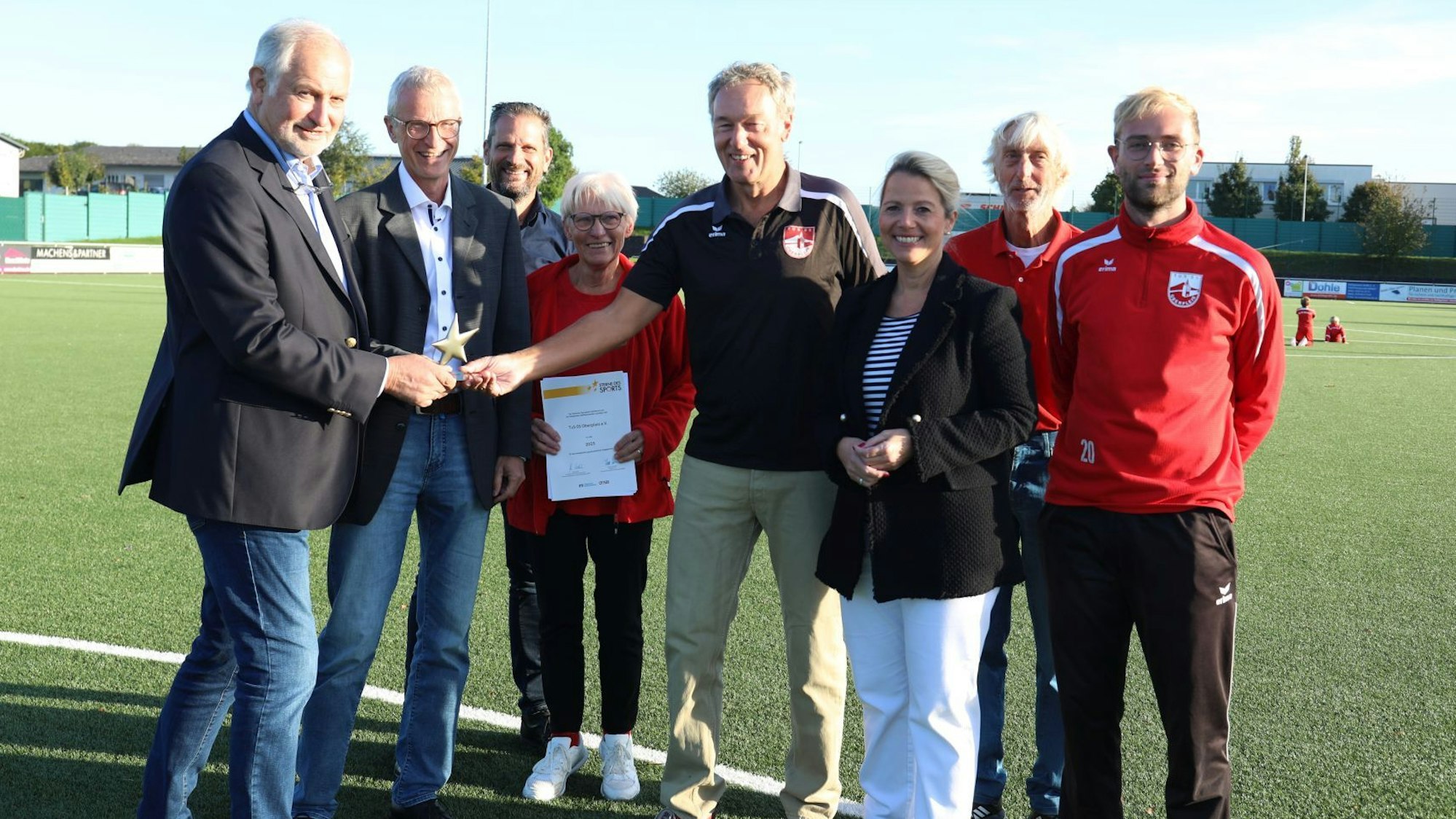 Eine Gruppe von Männern und Frauen steht auf dem Kunstrasenplatz des TuS 05 Oberpleis. Vorsitzender Norbert Seeger nimmt die Auszeichnung entgegen.