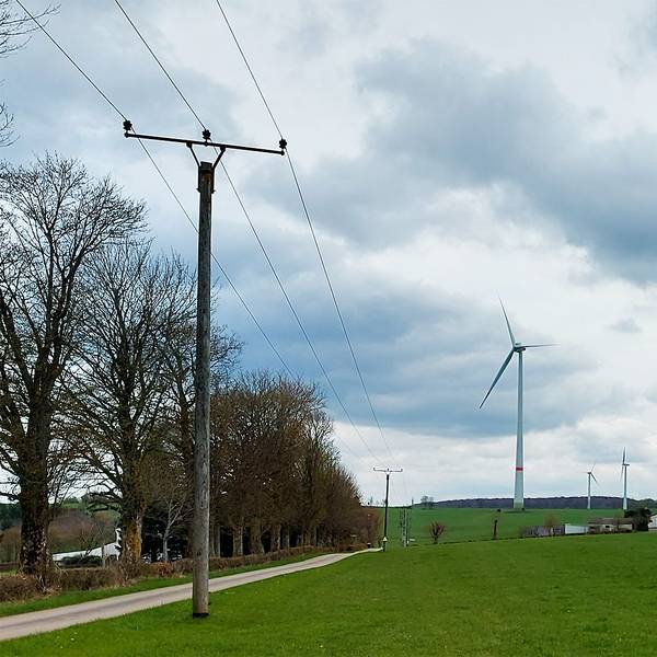 Auf einer Wiese sollen Photovoltaik-Module aufgebaut werden. Im Hintergrund sind ein Bauernhof und ein Windrad zu sehen, links eine Stromleitung.