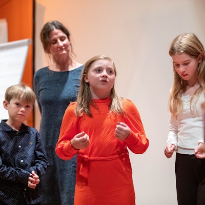 Ein Mädchen hält einen Vortrag, zwei Schulkinder und eine Frau hören zu.