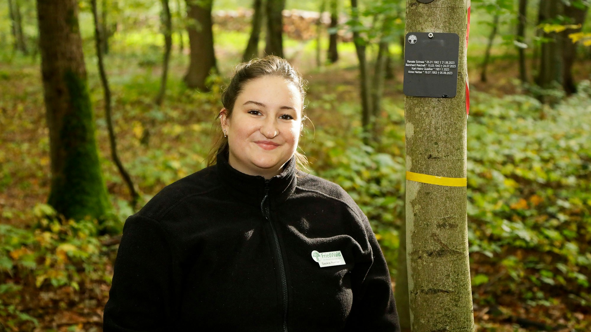 Die junge Frau in schwarzer Fleecejacke steht vor einem Baum mit Plakette der hier Bestatteten.