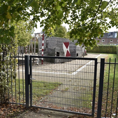 Hinter einem grünen Tor sind Holzspielgeräte eine Spielplatzes zu erkennen.