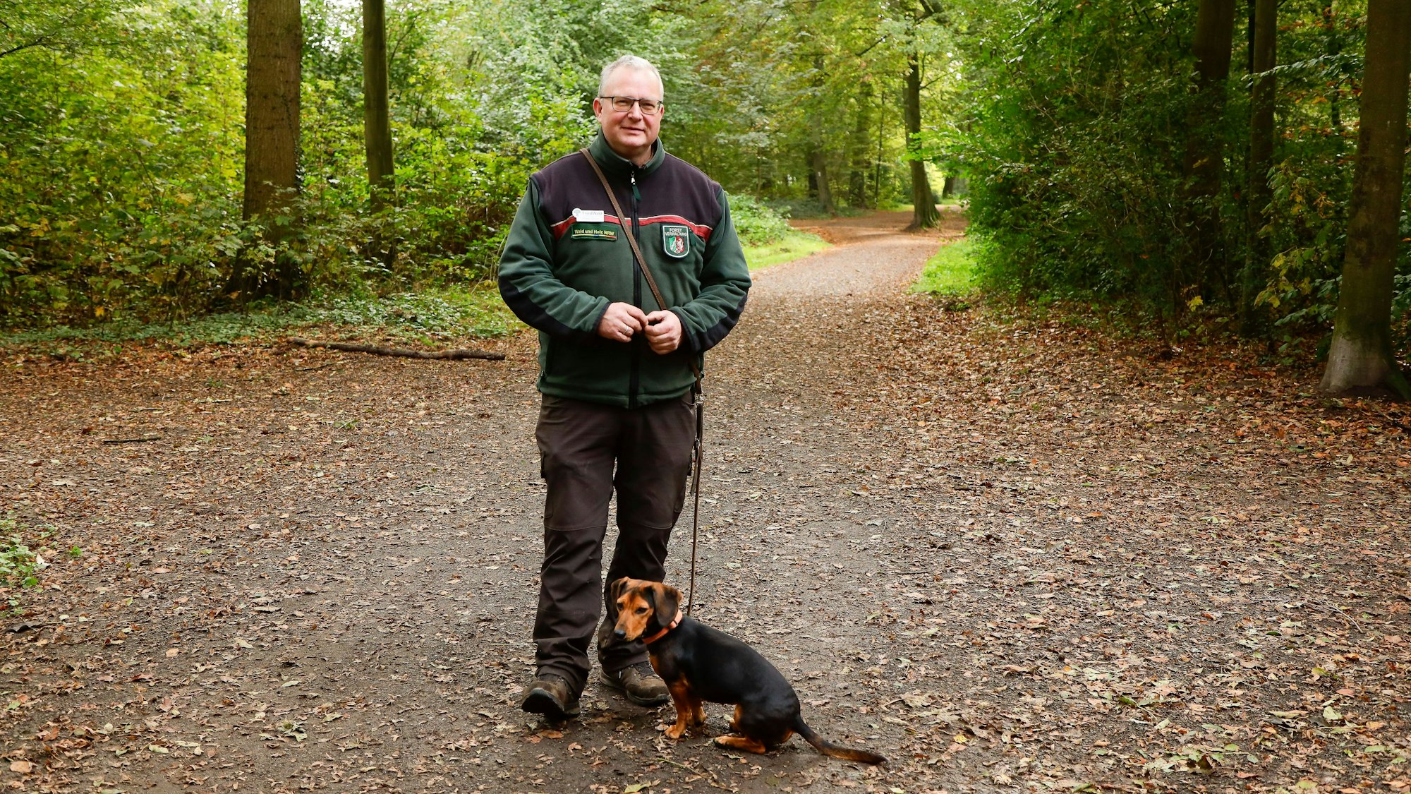 Der Förster steht in grüner Fleecejacke des Landesbetriebs auf einem Waldweg, die kleine Hündin an der Leine sitzt vor ihm.