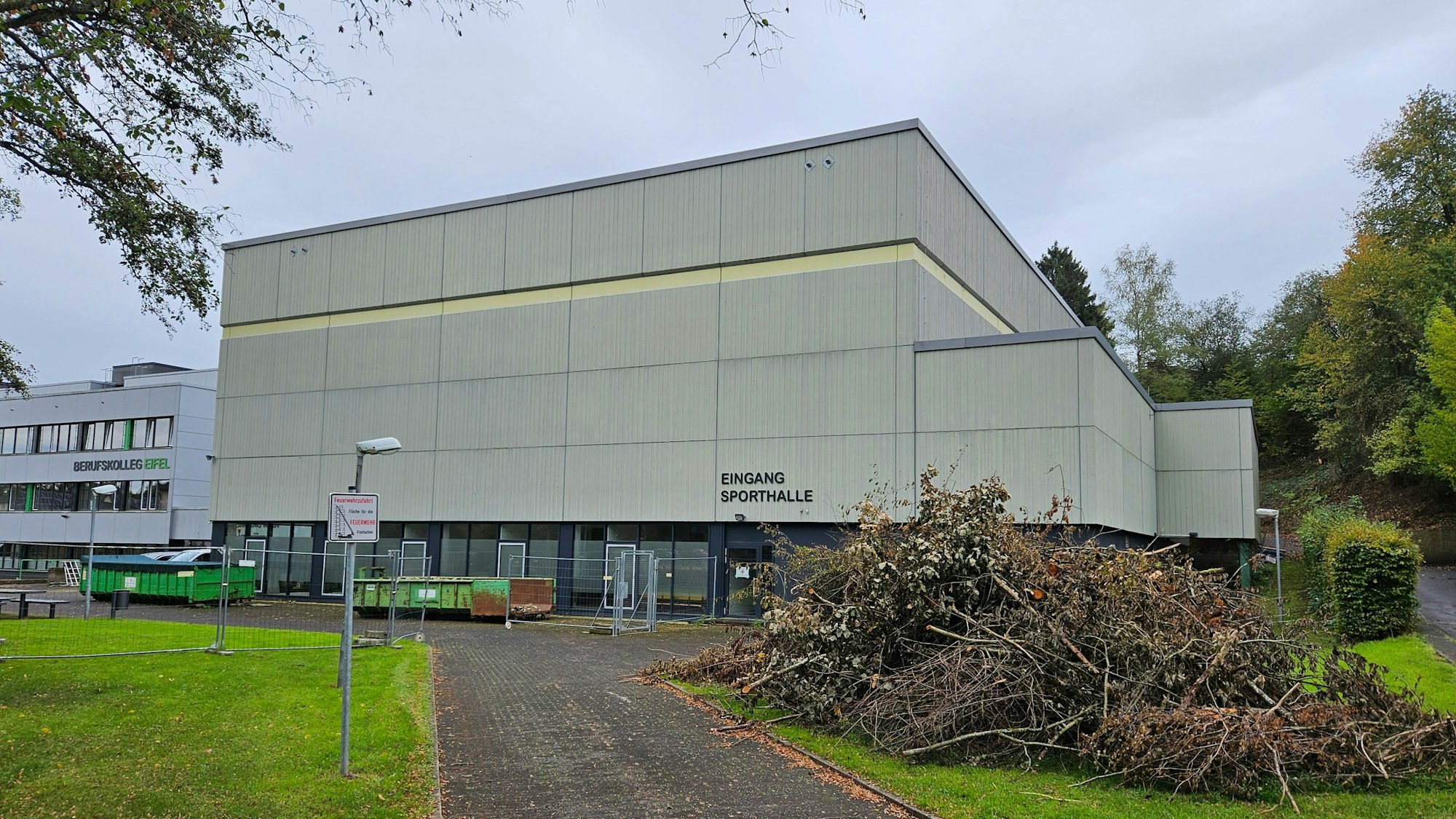 Zu sehen ist die Sporthalle Kall, vor der ein Haufen mit abgeschnittenen Ästen liegt.