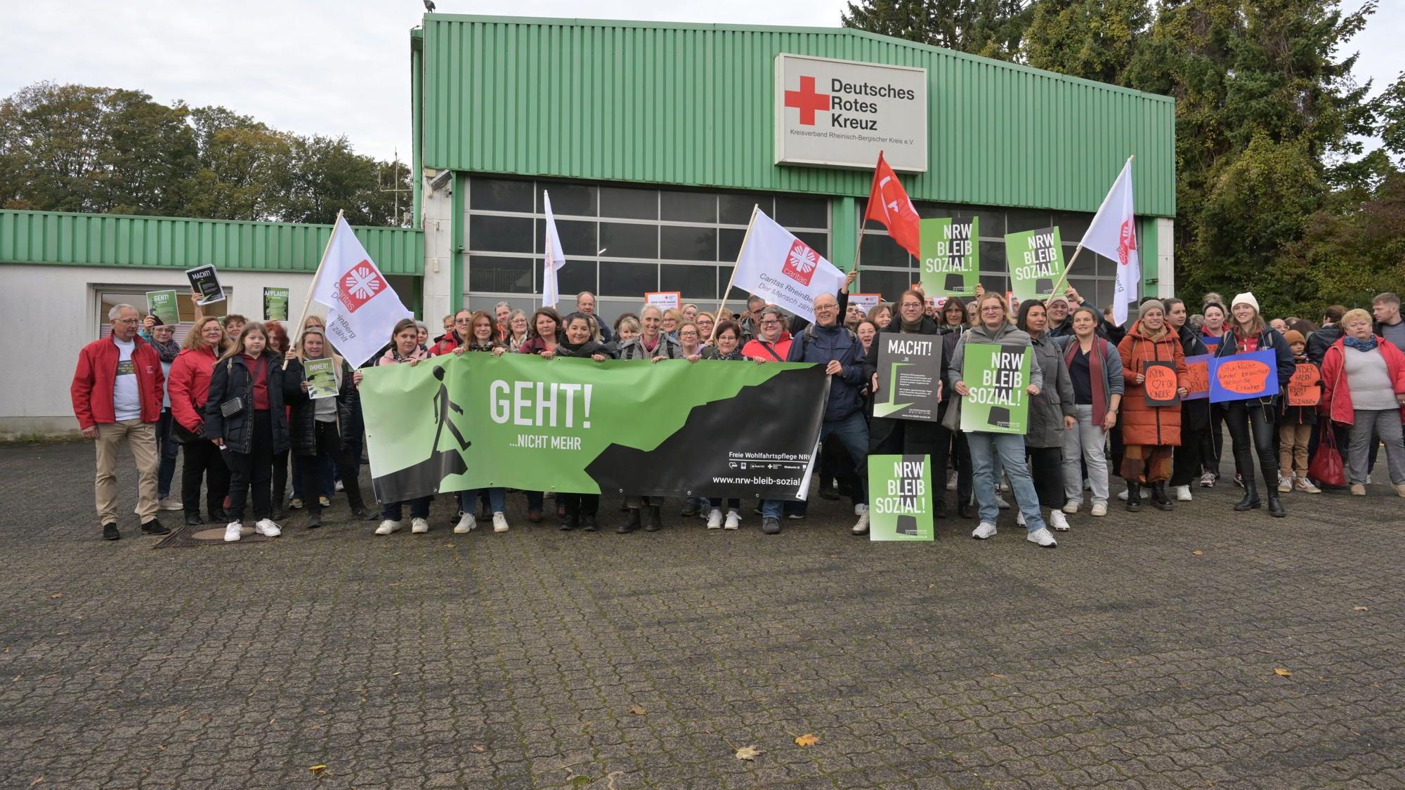 Vor einer Halle des DRK haben sich die Mitarbeiter versammelt und halten Plakate und Fahnen hoch.