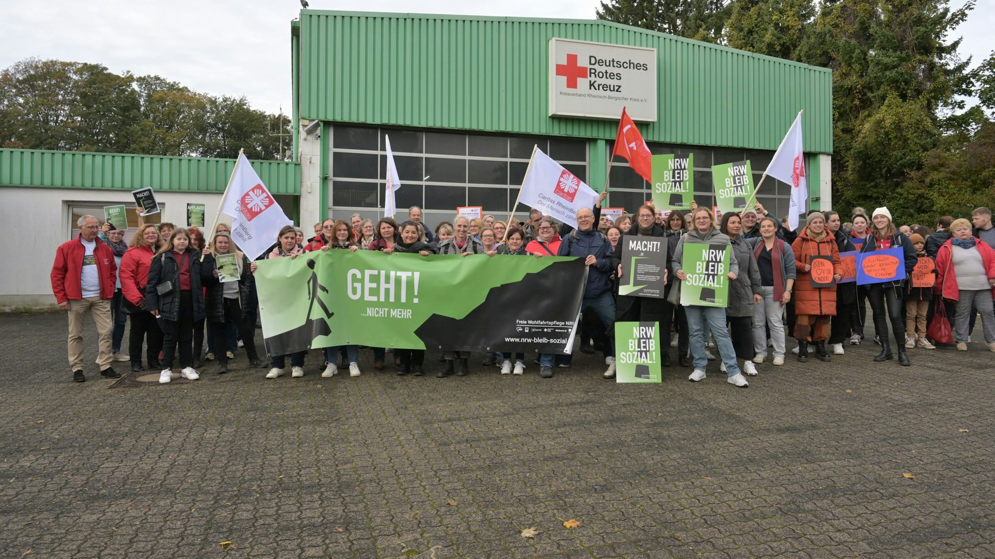 Vor einer Halle des DRK haben sich die Mitarbeiter versammelt und halten Plakate und Fahnen hoch.