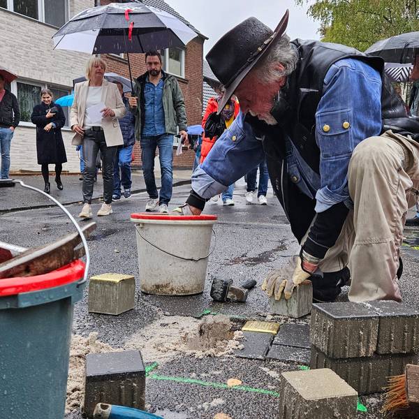 Ein Mann mit Hut kniet auf dem Gehweg und verlegt Pflasterstein, Menschen mit Regenschirmen beobachten ihn dabei.