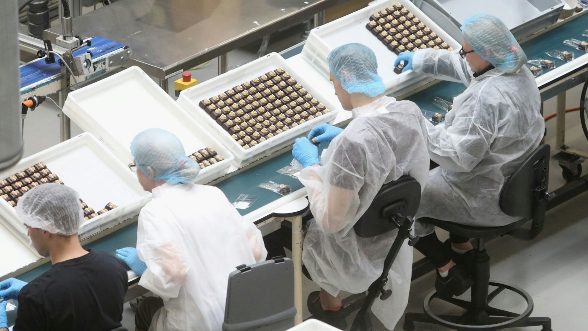 Zwei Männer und zwei Frauen mit Haarhauben und Kitteln sind in Handarbeit mit Pralinen beschäftigt.