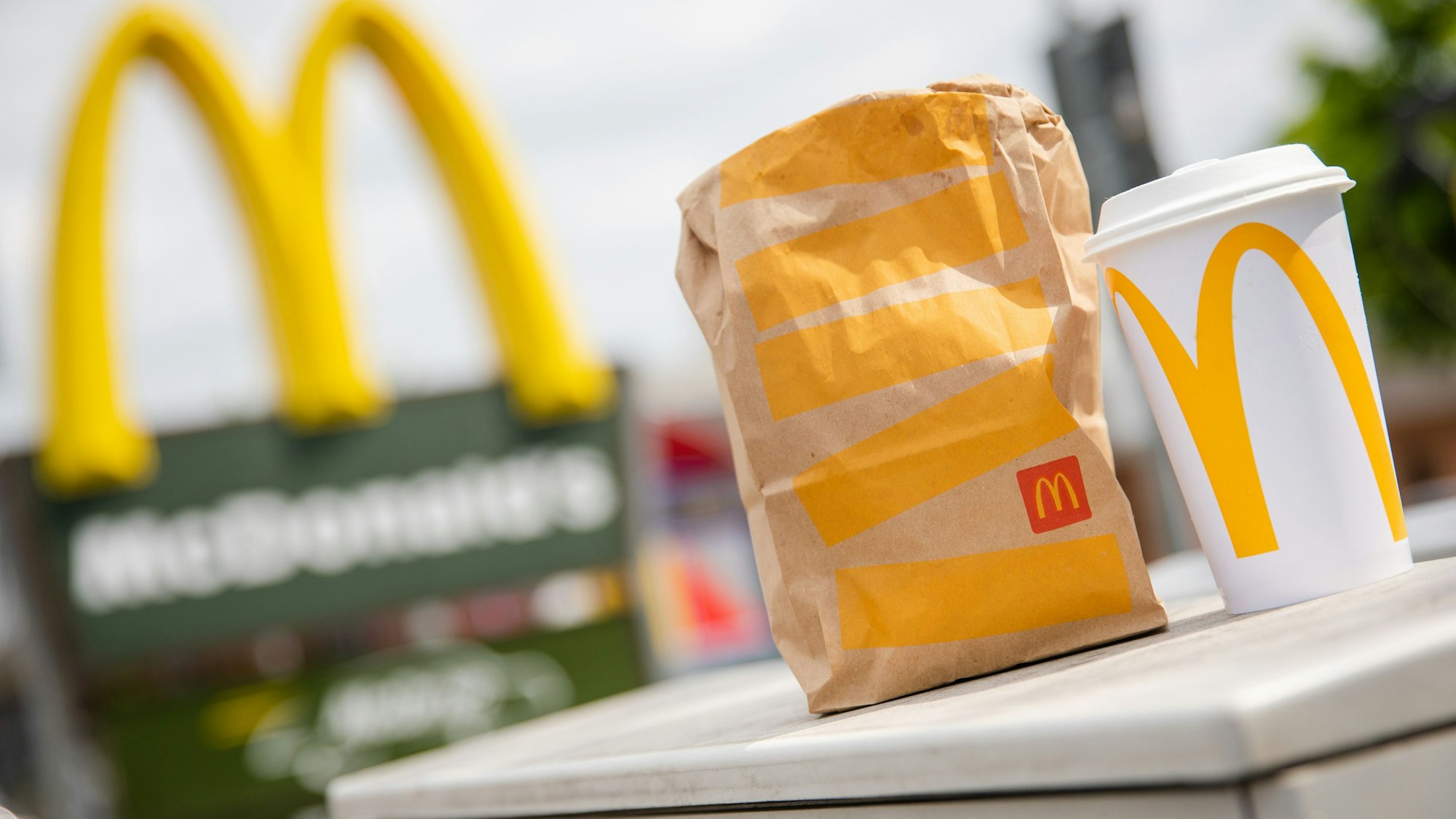 Eine McDonald’s To-Go-Tüte und ein To-Go-Becher stehen vor einer McDonald’s-Filiale.