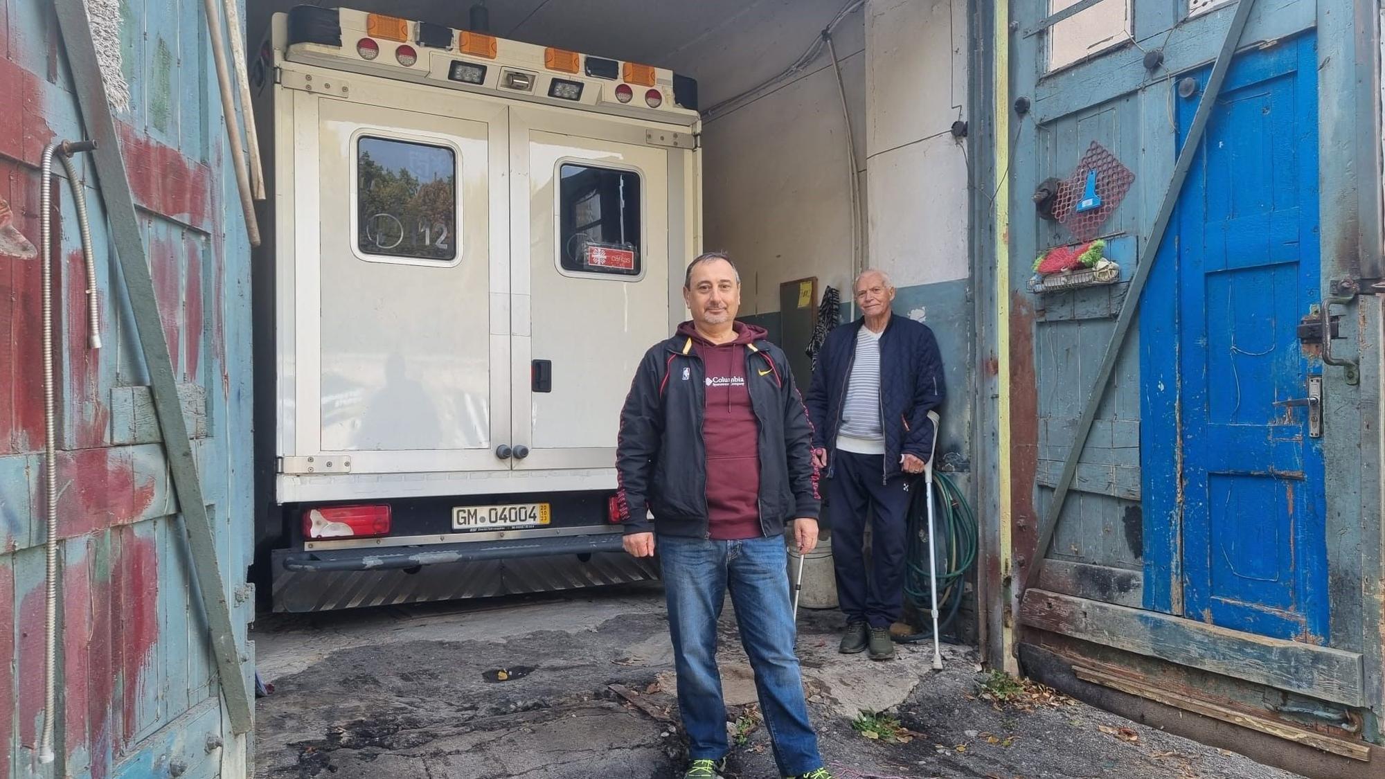 Ein Rettungswagen steht in einer Garage. Zwei Männer, einer mit Krücken, stehen davor.