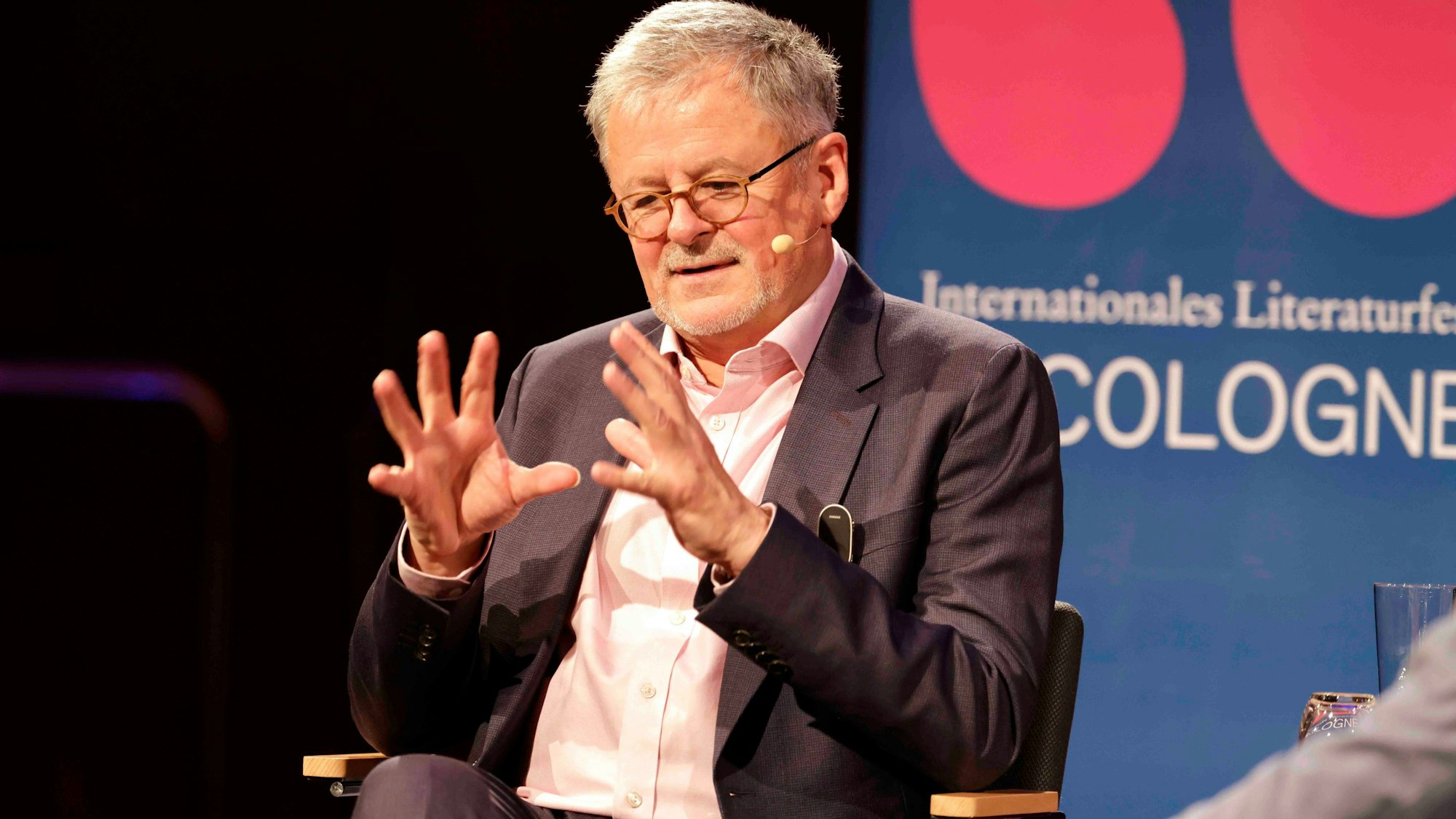 Sir Christopher Munro Clark, gekleidet in einen dunklen Anzug und ein weißes Hemd, sitzt gestikulierend in einem Stuhl und spricht.