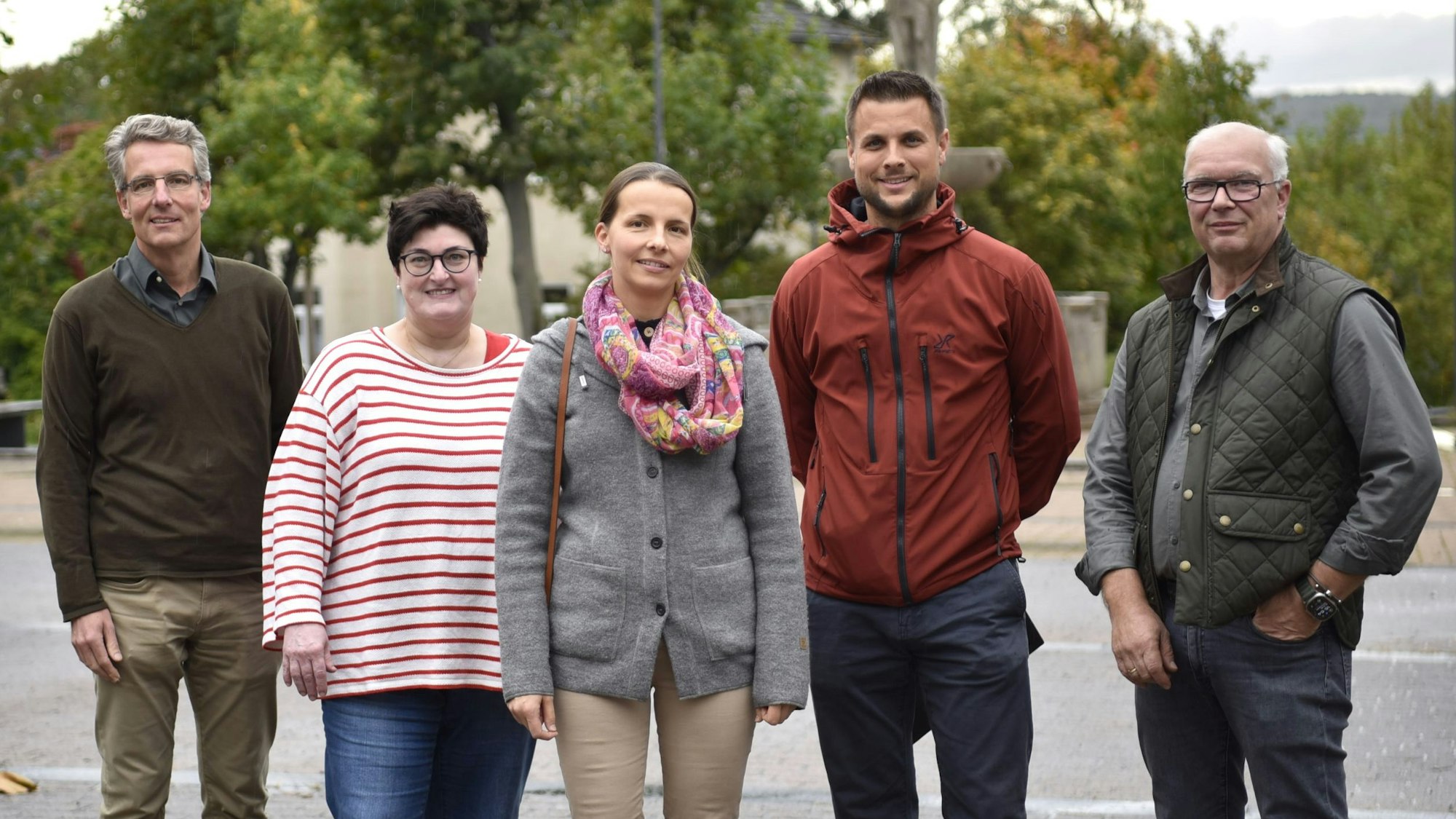 Drei Männer und zwei Frauen stehen auf einem Platz und lächeln in die Kamera, im Hintergrund sind Bäume zu erkennen.