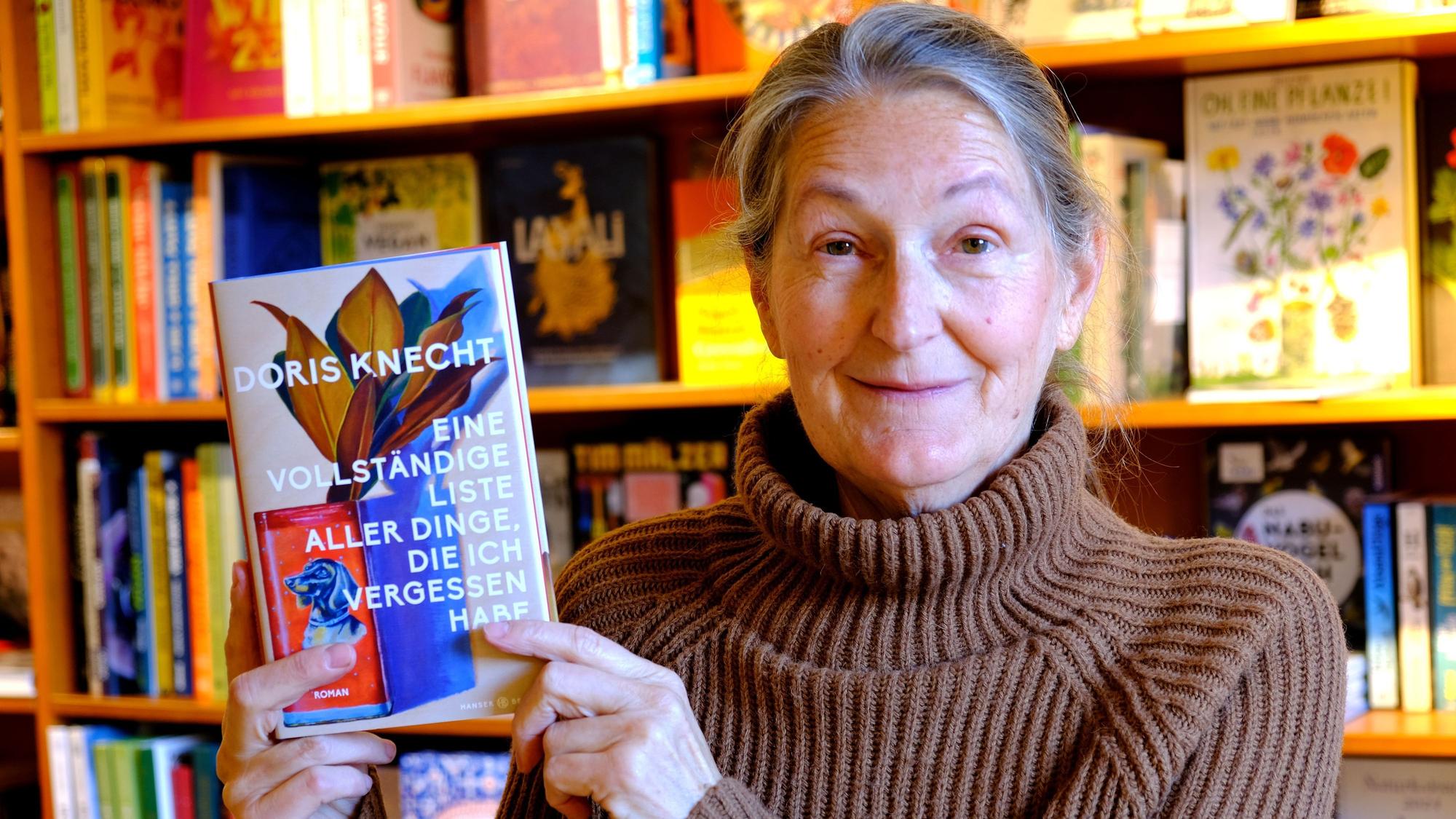 Iris Pütz steht vor einem Buchregal. Sie hat ein Exemplar eines Buchs von Doris Knecht in der Hand.
