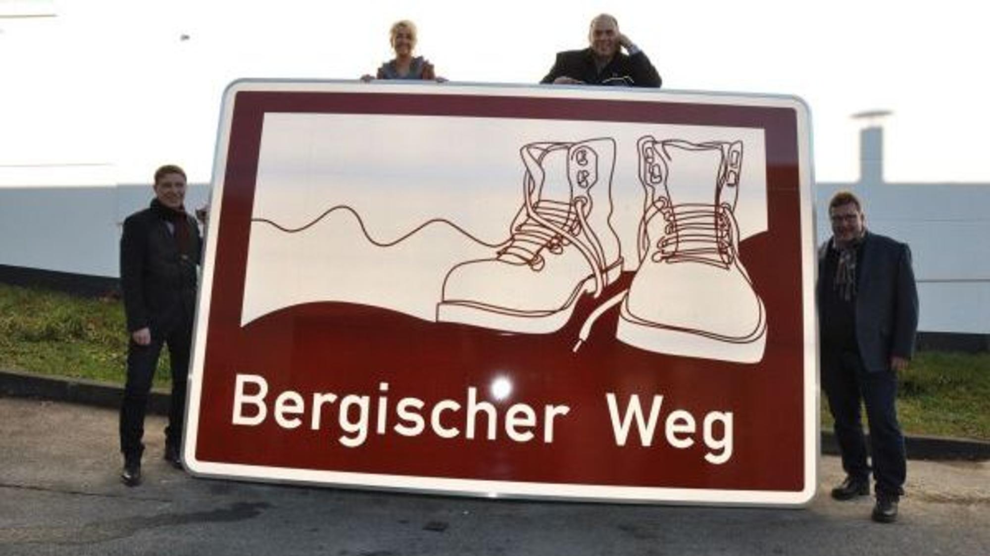 Vier Menschen stehen an einem Schild, auf dem Wanderschuhe und der Schriftzug „Bergischer Weg“zu sehen sind.