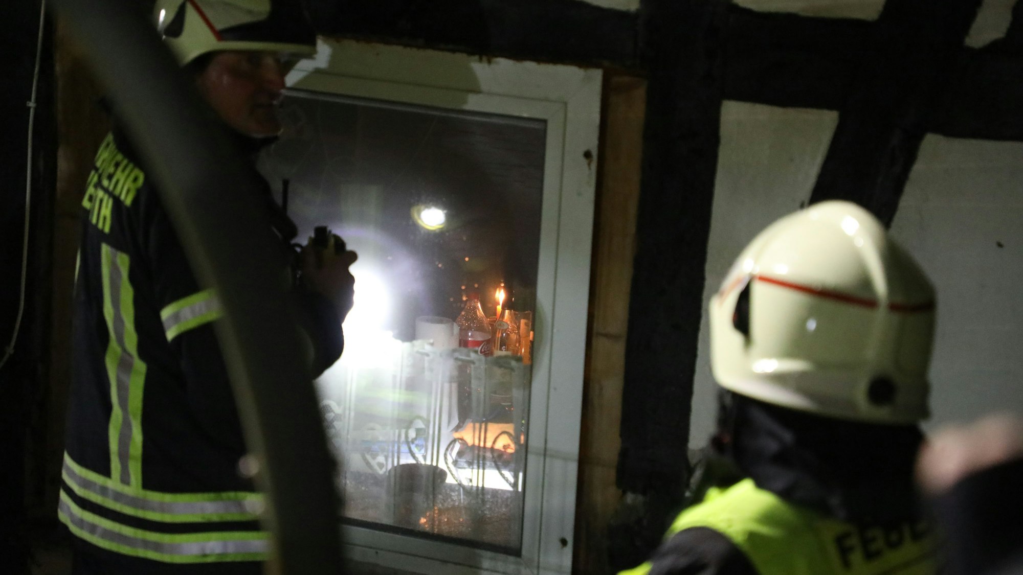Ein Feuerwehrmann leuchtet mit einer Taschenlampe durch ein Fenster in einen Raum eines Fachwerkhauses, in dem Kerzen auf dem Tisch brennen.