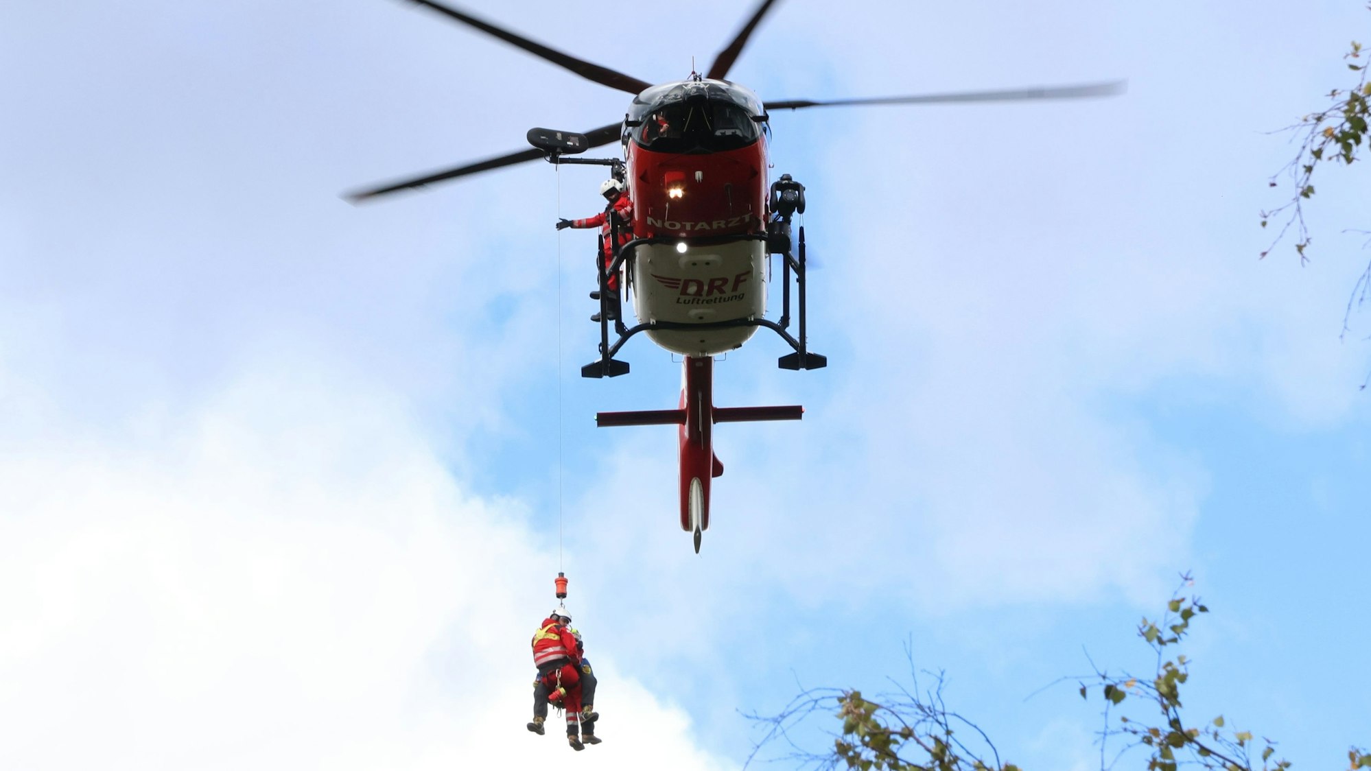 Zu sehen ist ein Rettungshubschrauber, der zwei Menschen mit einer Winde nach oben zieht.