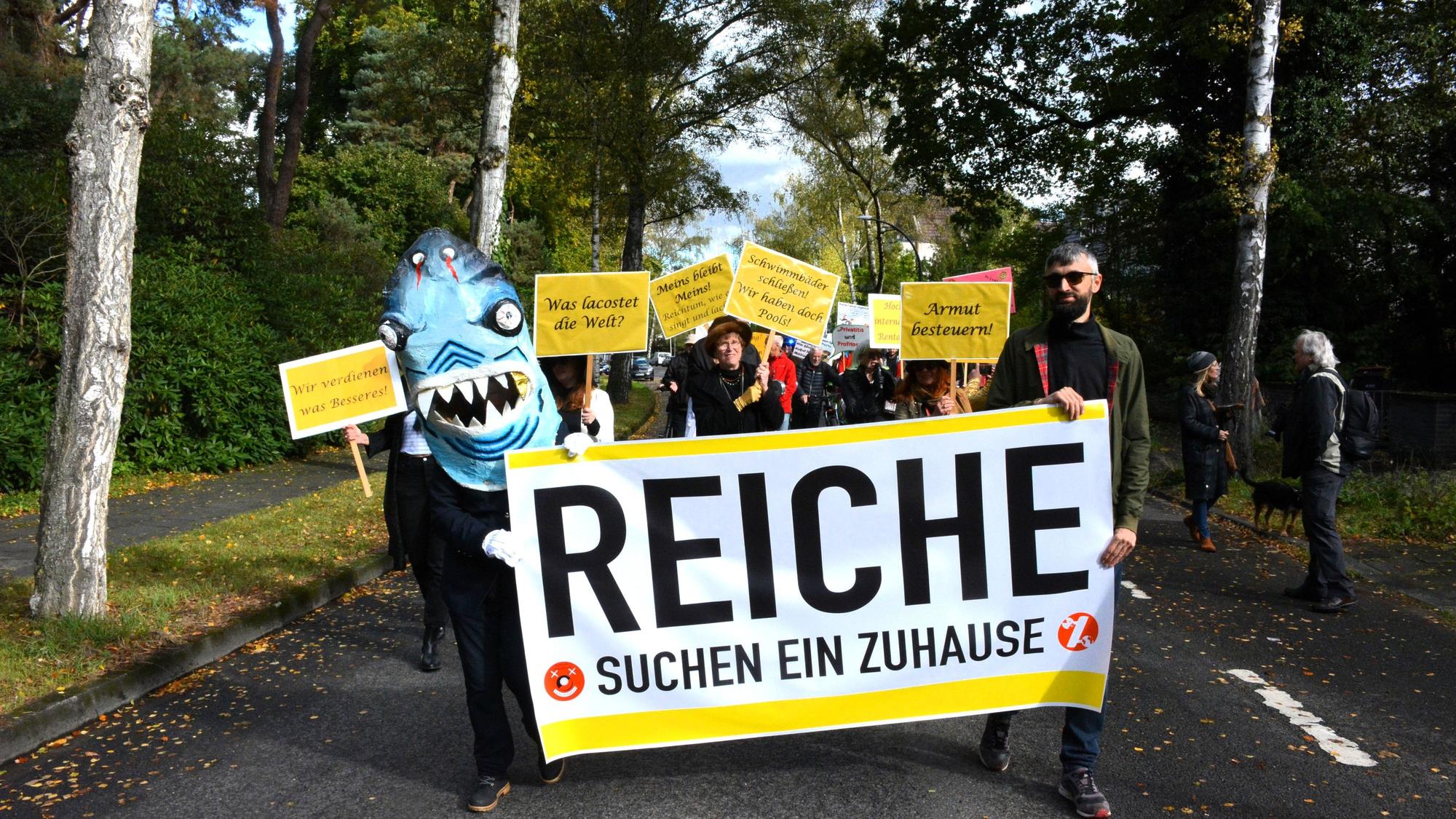 Ein als Miethai maskierter Demonstrant und ein Mann tragen ein Transparent, auf dem „Reiche suchen ein Zuhause“ steht.