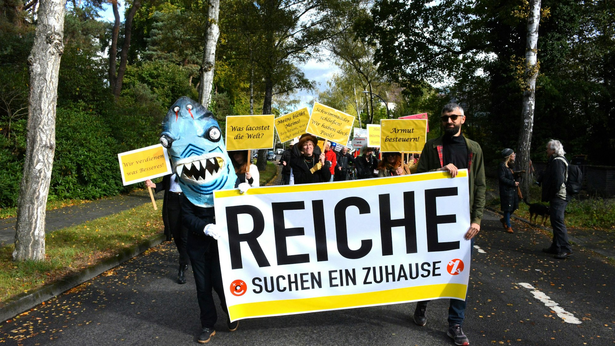 Ein als Miethai maskierter Demonstrant und ein Mann tragen ein Transparent, auf dem „Reiche suchen ein Zuhause“ steht.