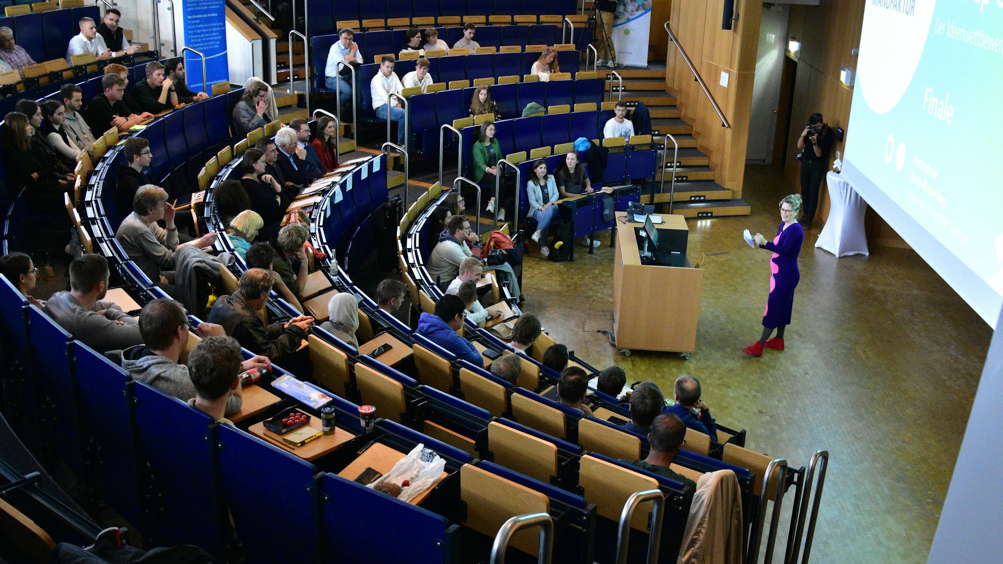 Voller Hörsaal, Frau in buntem Kleid moderiert