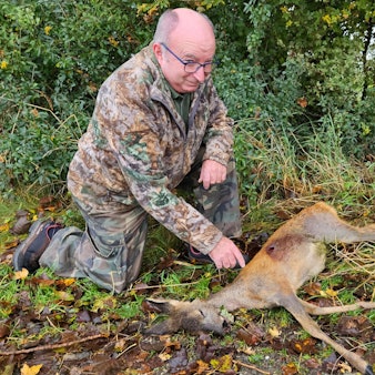Ein Mann in Tarnkleidung kniet neben einem toten Reh, das eine Wunde hat.