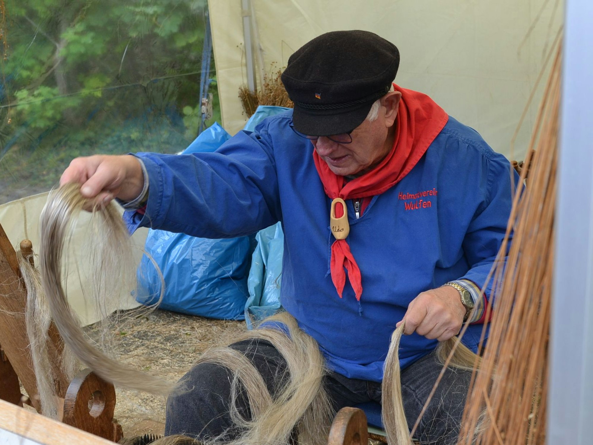 Ein Mann in einem blauen Hemd und mit einem roten Tuch um den Hals sitzt auf einem Stuhl. Er verarbeitet Flachs zu Leinen.