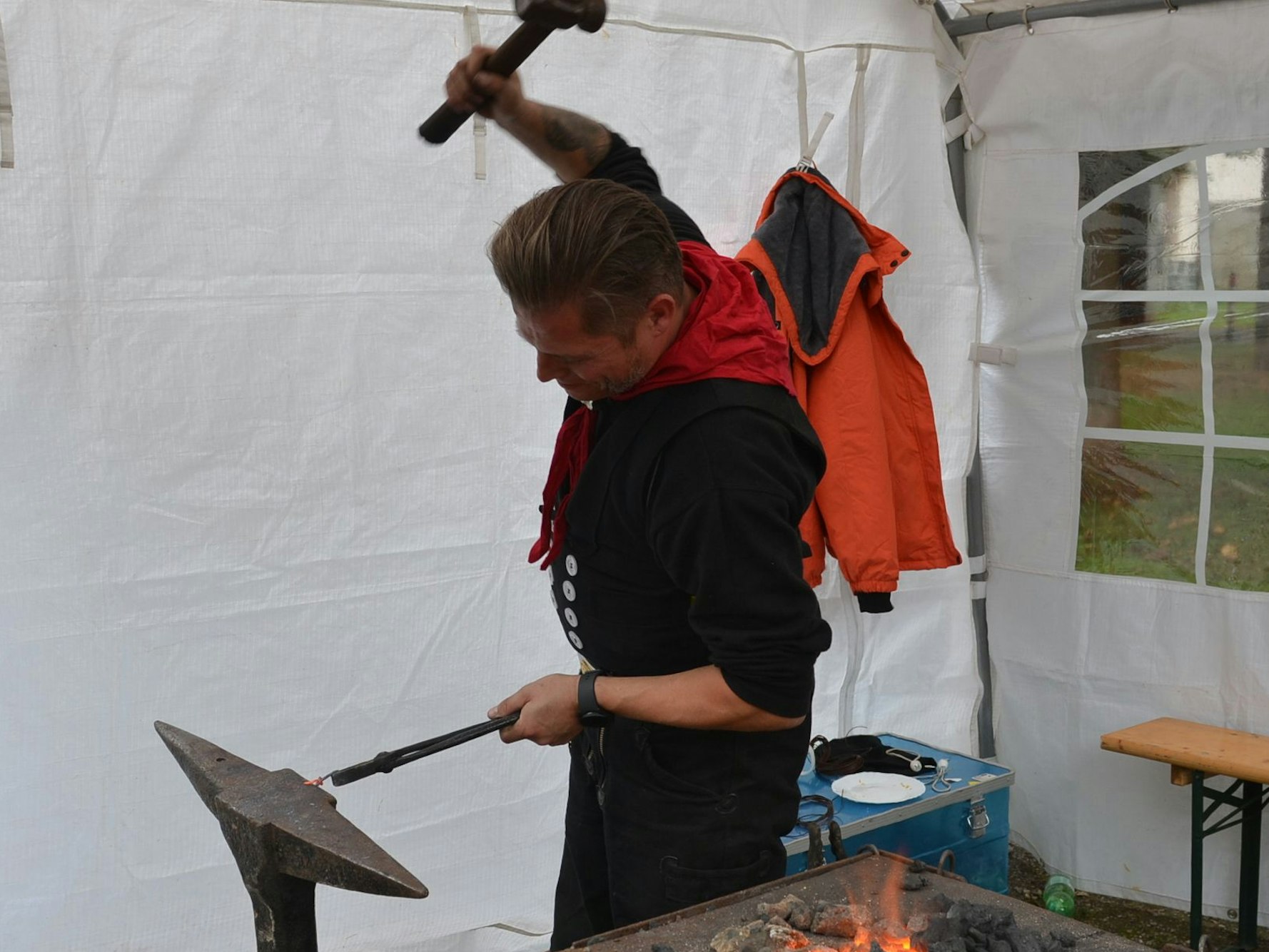 Schmied Patrick Duwenbeck holt mit dem Hammer in seiner rechten Hand aus, um ihn auf ein rotglühendes Metall zu schlagen, das er mit der linken auf einem Amboss festhält. Das Schmiedefeuer ist neben ihm zu sehen.