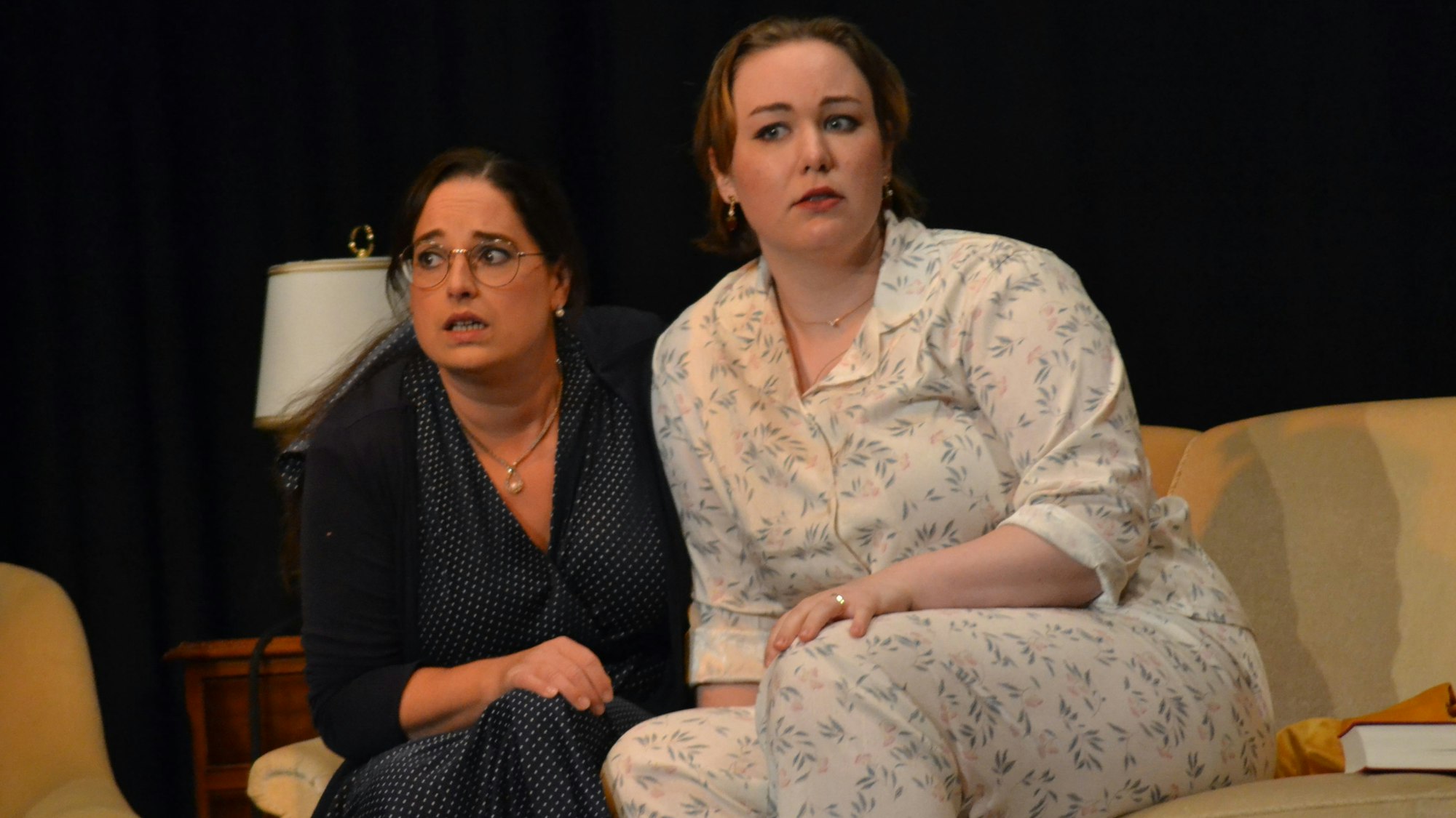 Zwei Frauen sitzen auf einem hellen Sofa; eine trägt einen Schlafanzug, die andere ein dunkles Kleid.