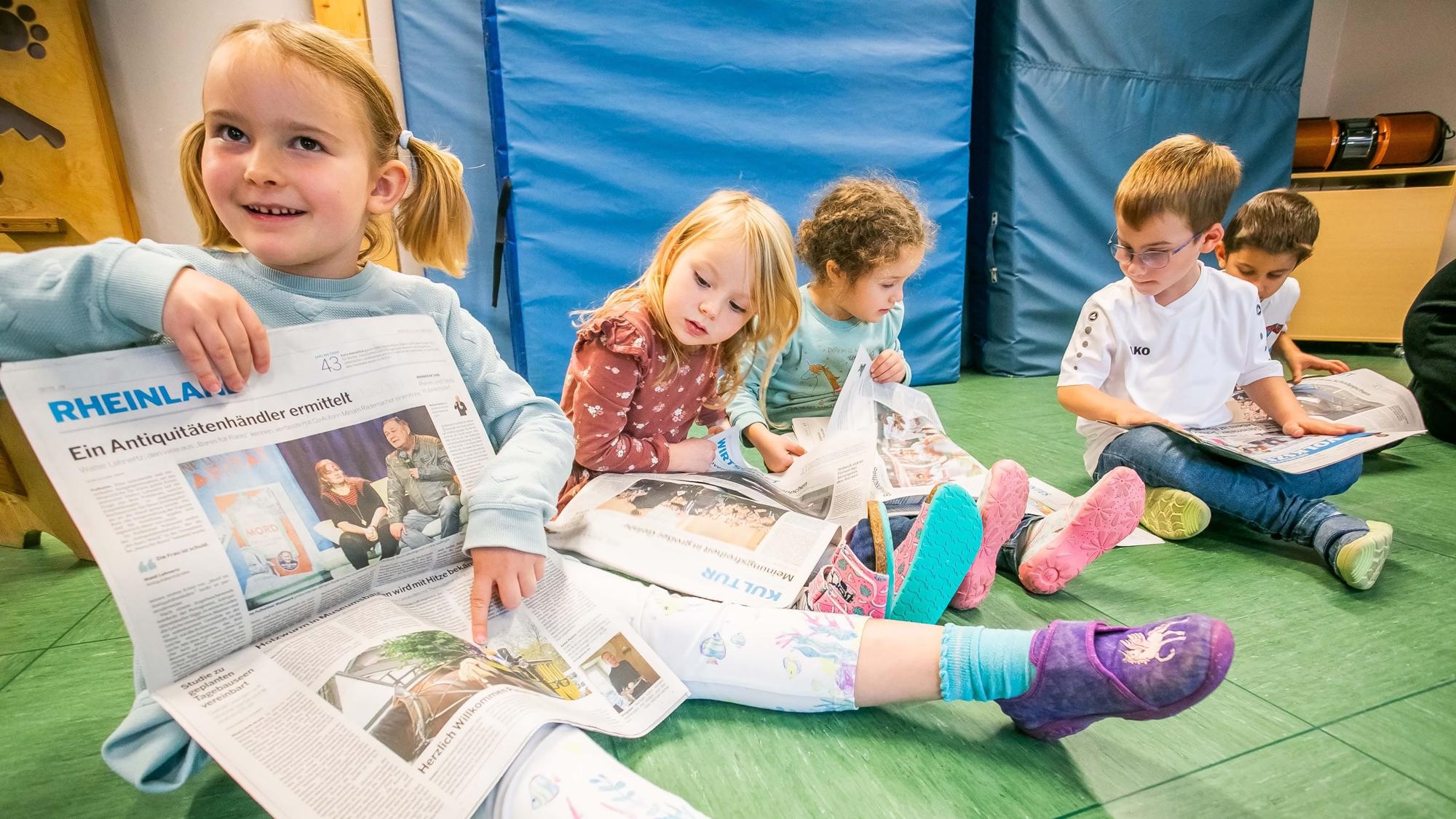 Fünf Kinder sitzen auf dem Boden und schauen sich Tageszeitungen an.