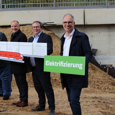 Bernd Kolvenbach (Vorsitzender der Verbandsversammlung von go.Rheinland), Dr. Marcus Schenkel (Leiter Infrastrukturprojekte West der DB), NRW-Verkehrsminister Oliver Krischer und Dr. Norbert Reinkober stehen vor der Baustelle einer Brücke und halten Tafeln mit den Aufschriften„ Startschuss“und „Elektrifizierung“ sowie mit einem gemalten Zug und einer gemalten Zugstrecke samt Stromleitung in den Händen.