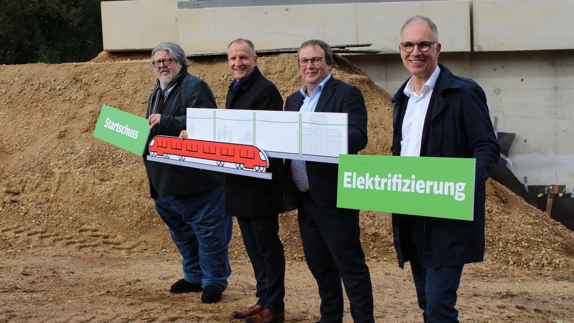 Bernd Kolvenbach (Vorsitzender der Verbandsversammlung von go.Rheinland), Dr. Marcus Schenkel (Leiter Infrastrukturprojekte West der DB), NRW-Verkehrsminister Oliver Krischer und Dr. Norbert Reinkober stehen vor der Baustelle einer Brücke und halten Tafeln mit den Aufschriften„ Startschuss“und „Elektrifizierung“ sowie mit einem gemalten Zug und einer gemalten Zugstrecke samt Stromleitung in den Händen.